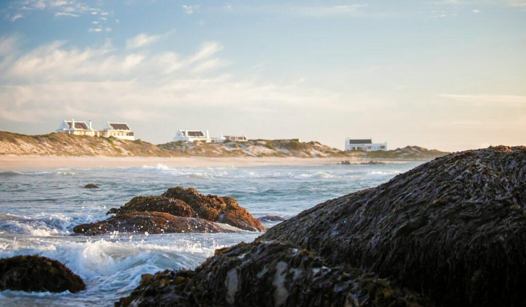 Holiday homes on beach front at sunset with crashing waves on boulders in the foreground.