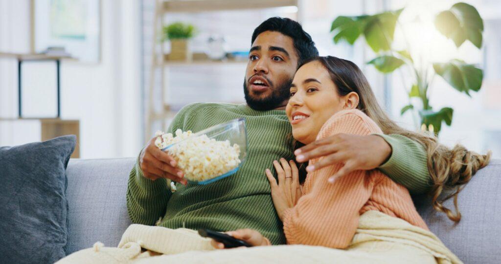 Jump scare, hug and couple watching tv on a sofa with popcorn for movie, film or streaming show at