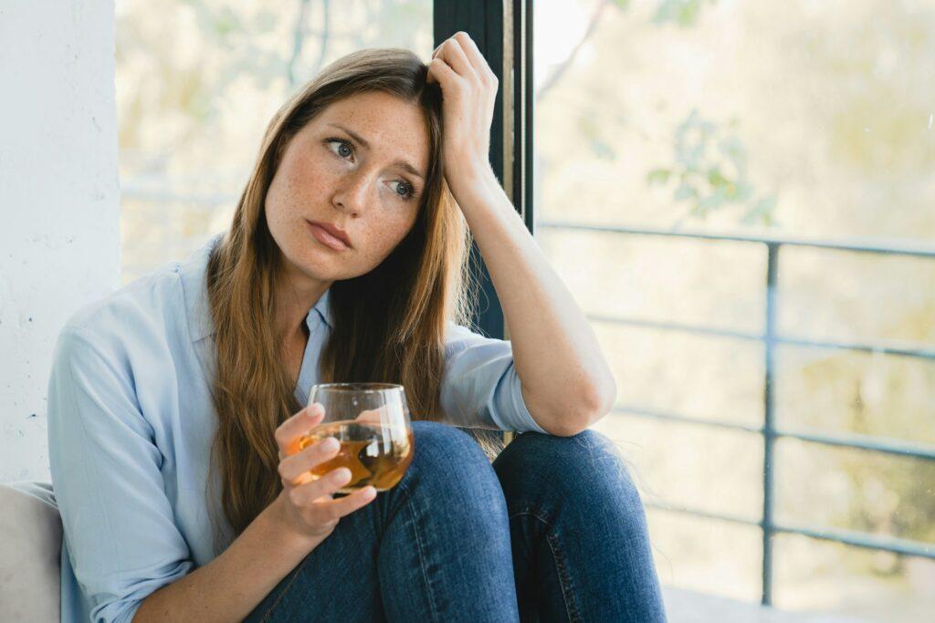 Sad stressed young woman thinking about bad memories drinking juice by the window alone.