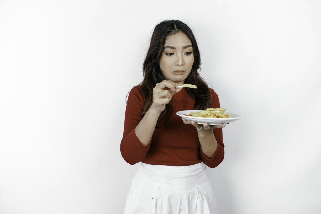 Young disgusted woman in casual sweater is holding french fries potato with disgusted gesture.