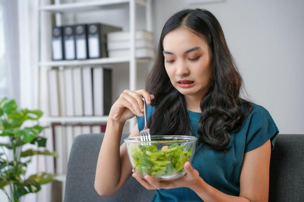 Young woman making disgusted face while eating salad at home