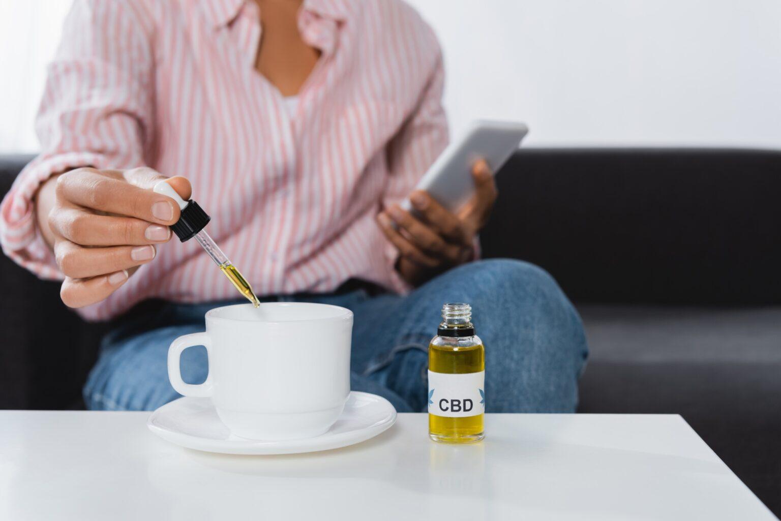 cropped view of african american woman holding smartphone and pipette with cbd liquid near cup of