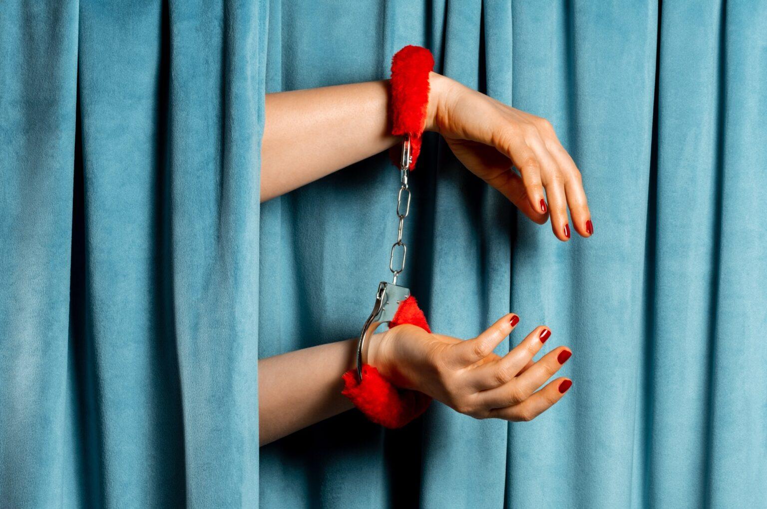 Female hand with red fluffy handcuffs on blue velvet curtains background.