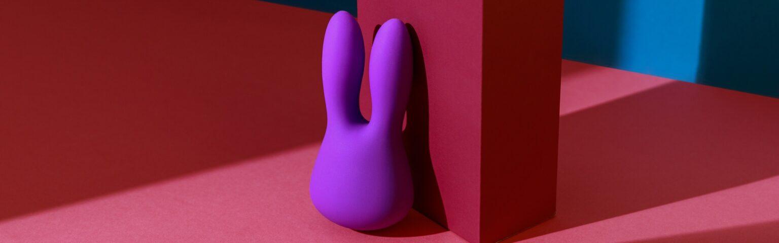 Comment choisir le bon accessoire pour pimenter sa vie intime ? Violet dildo vibrator for clitoris with pink gift box on colored background. Sex toy for