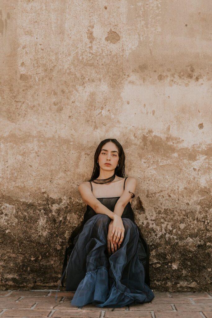 A woman in an elegant blue gown sits against a weathered vintage wall, exuding grace and serenity.
