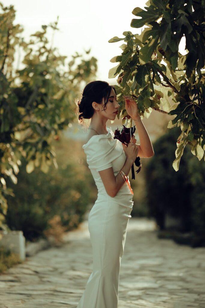 Comprendre la dépression saisonnière : symptômes et solutions Elegant woman in a white dress stands in a sunlit garden, touching leaves.