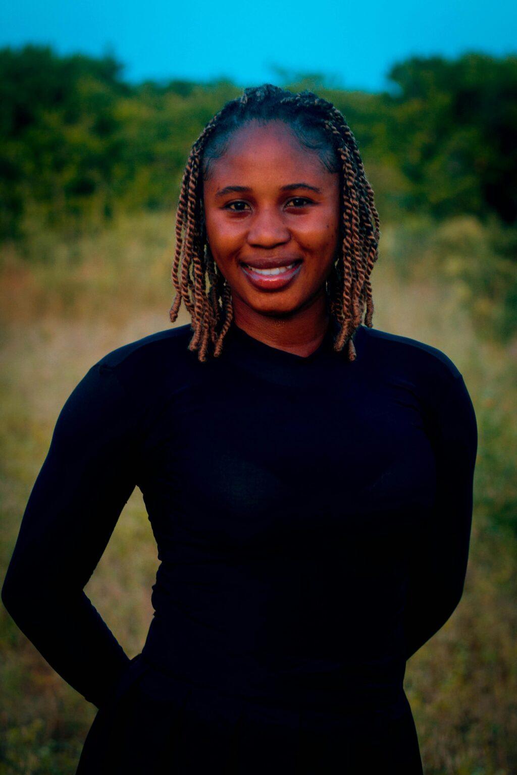 A woman smiles warmly while standing outdoors in a natural setting, surrounded by greenery.
