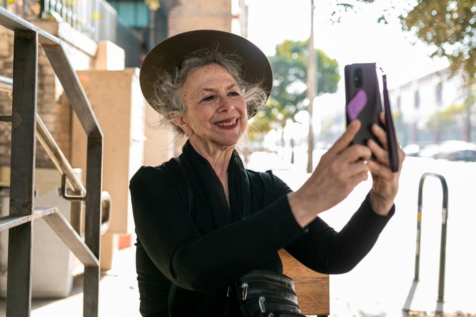 Senior woman in black attire taking a selfie on a sunny day, embracing modern technology.