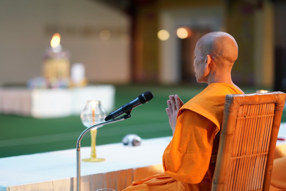 Le rôle de la religion et de la spiritualité dans le but de vie A Buddhist monk in orange robes sits meditatively at a table with a microphone in a temple.