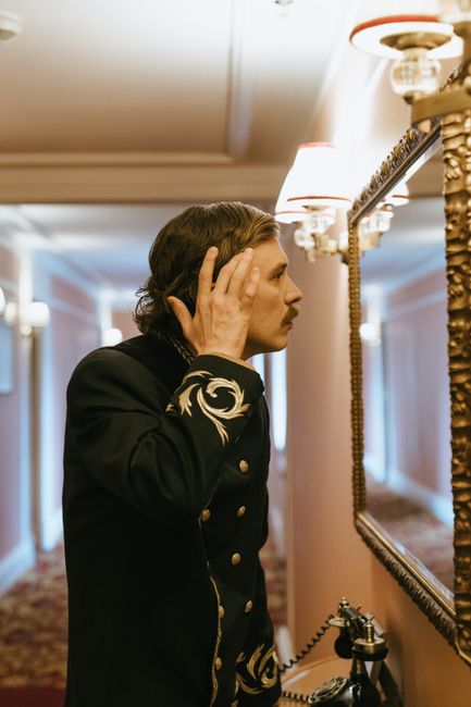 A bellboy in a stylish uniform checks his appearance in a hotel corridor mirror.