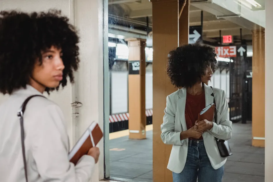 A fashionable woman with curly hair and a notebook reflected in a subway mirror.