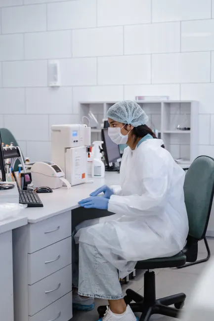 A female lab technician wearing protective gear sits at a desk in a clinical laboratory.