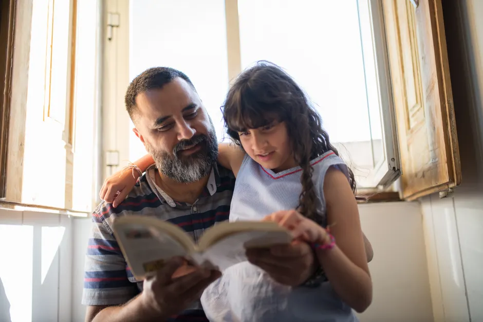 A loving father reads with his daughter by a sunlit window, fostering family bonding.