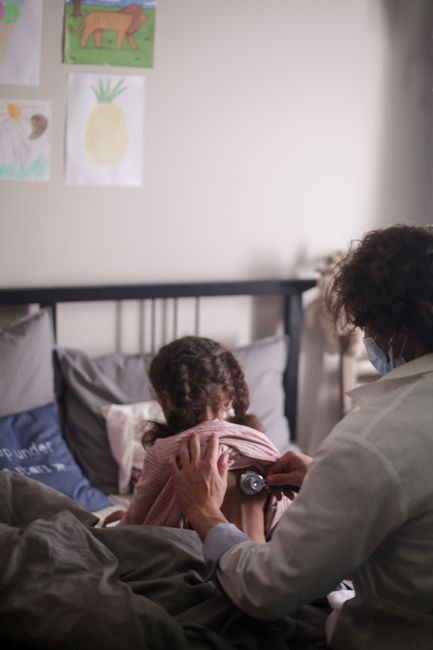 A pediatrician uses a stethoscope to examine a child sitting in a cozy bedroom.
