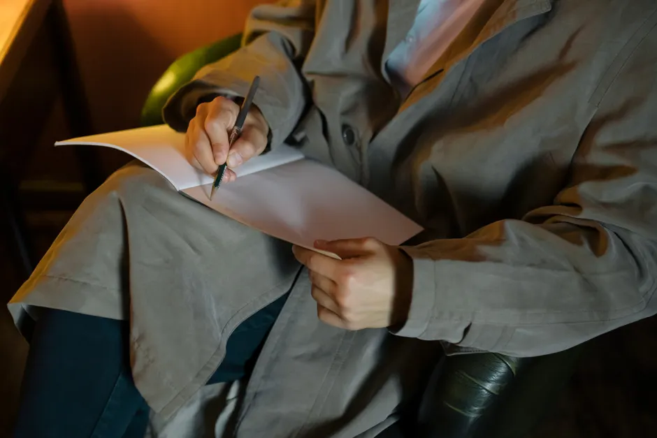 A person in a coat writes in a notebook in a warm, indoor environment, focused on creativity and thought.