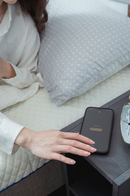 A person in bed reaches to snooze their smartphone alarm on a bedside table.
