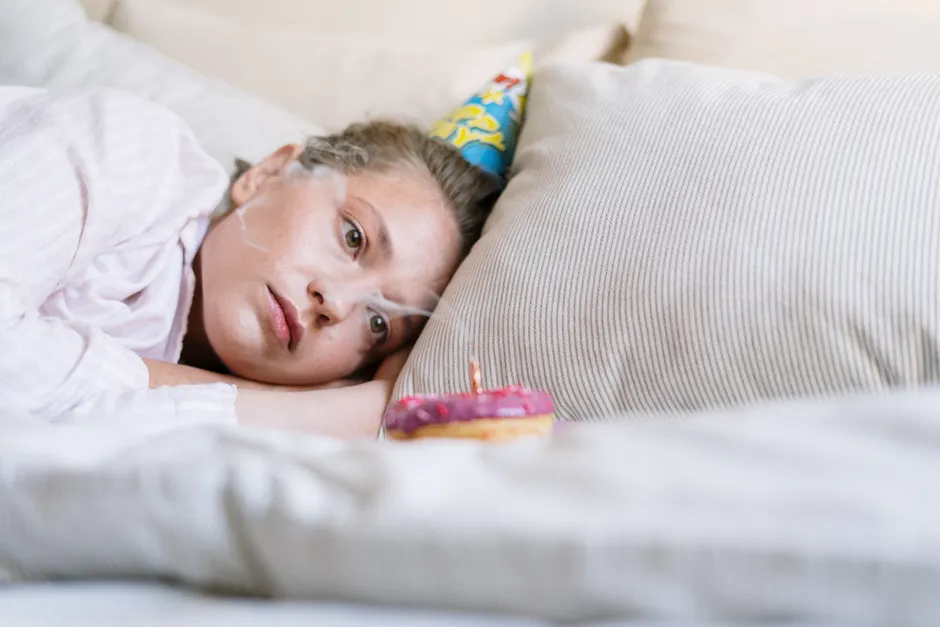 Partir tôt d’une soirée : pourquoi ce n’est pas forcément antisocial A person in pajamas lays in bed with a party hat and donut, reflecting on a quiet birthday.