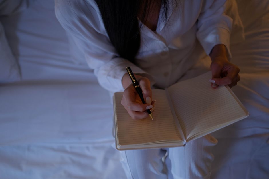 Défusion cognitive : prendre de la distance avec ses pensées négatives A person is writing in a notebook while sitting on a bed in a dimly lit room.
