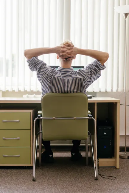 A person sitting at a desk in an office, viewed from behind with hands behind head.