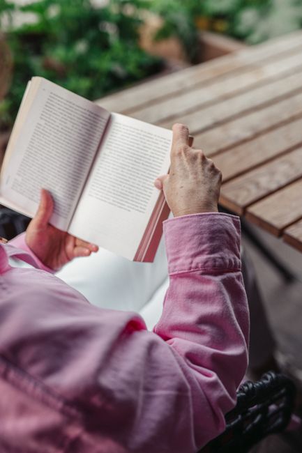 A person wearing a pink jacket is immersed in reading a book outdoors on a wooden table.