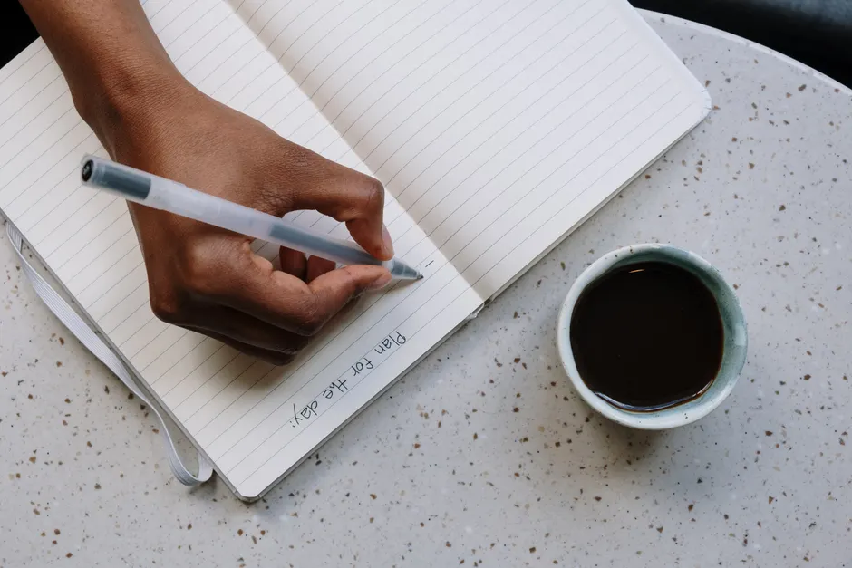 A person writes plans in a notebook beside a cup of coffee on a speckled table.