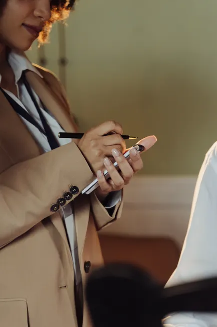 A professional adult woman in a beige blazer takes notes with a pen and notepad indoors.