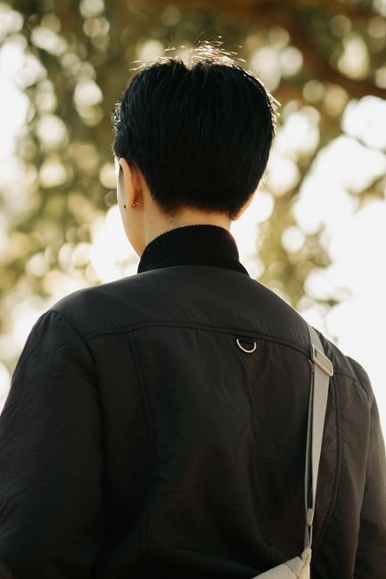 A stylish person with short hair wearing a black jacket, captured from behind against a natural background.