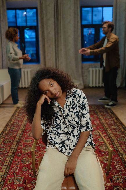 A teenager sits pensively on a chair amidst family conflict at home.