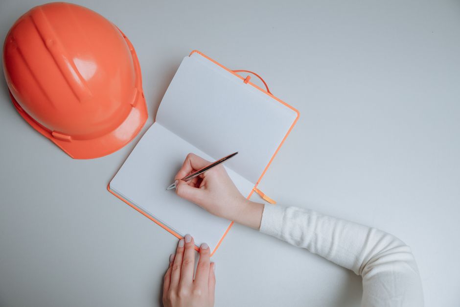 Rétablissement après l’automutilation : pourquoi il n’est pas linéaire A top view of a person writing in a notebook next to an orange hardhat on a white background.