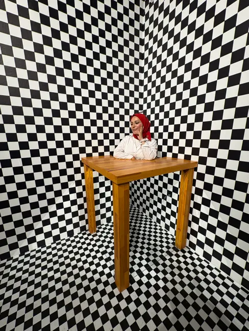 A woman sitting at a wooden table surrounded by a checkered pattern room creating an optical illusion.