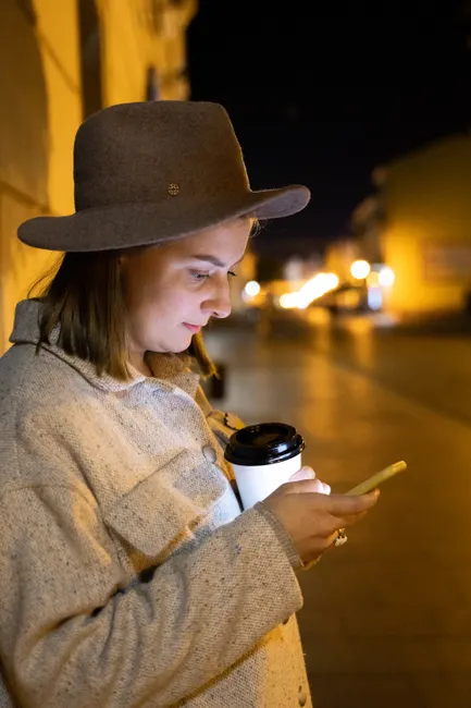 A woman wearing a hat uses her smartphone while holding a coffee cup on a city street at night.