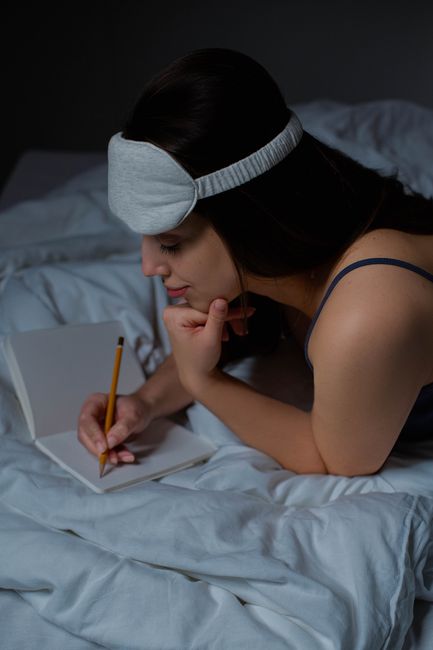 A woman wearing a sleep mask writes in a notebook while lying in bed.