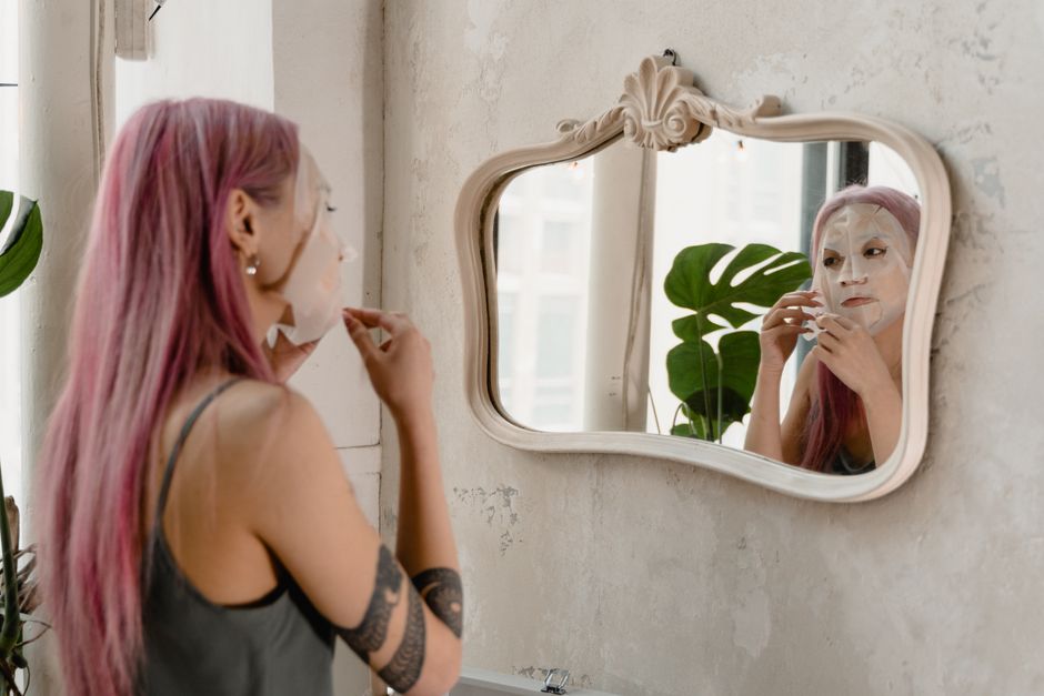 A woman with pink hair applies a facial mask in front of a mirror in a stylish room.