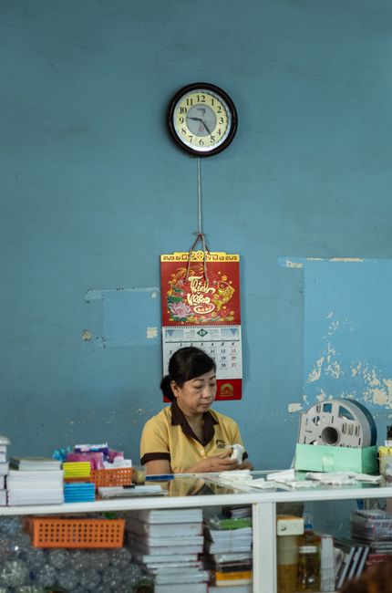 A woman working at a desk in a simple office with a clock on the wall and various office supplies.