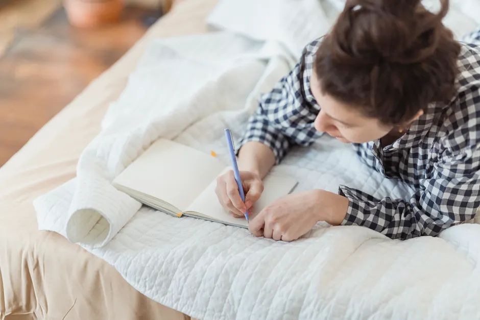 A woman writes in a journal while relaxing on a cozy bed with soft lighting and peaceful surroundings.