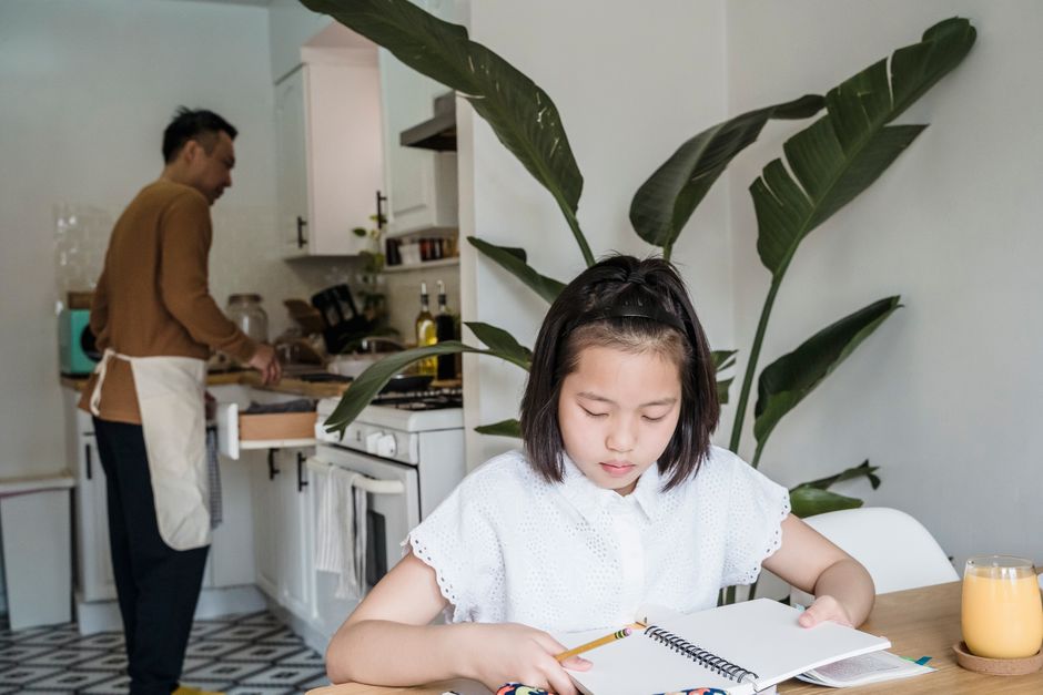 A young girl focuses on her homework while her parent cooks in a cozy kitchen setting.