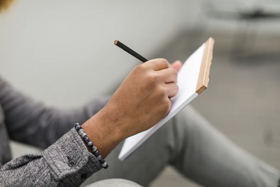 Trouble bipolaire et estime de soi : sortir du verdict permanent An adult hand writes in a notebook with a pencil, wearing a beaded bracelet and gray plaid sleeves.