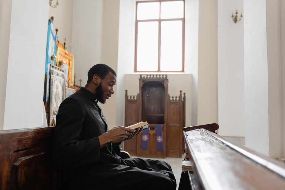 Traumatisme religieux : quand la foi devient une source d’angoisse An African American priest sitting in a church pew, reading a book in a serene church interior.