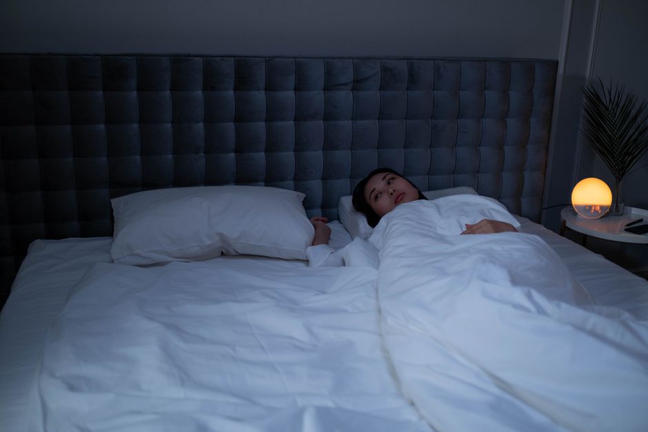 An Asian woman lying awake under soft white bedding in a dimly lit modern bedroom.