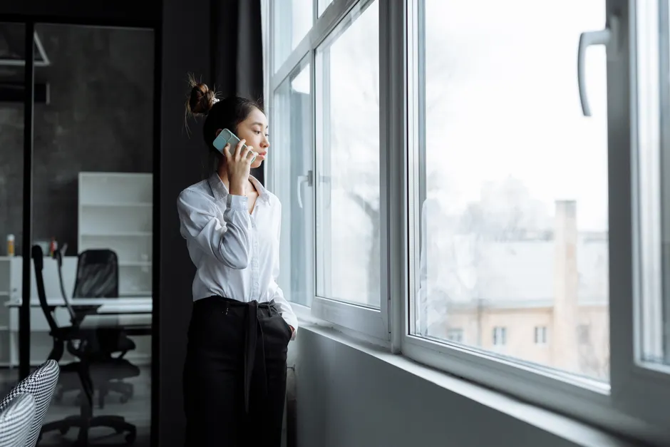 Asian woman in office clothes making a phone call while gazing outside a large window.