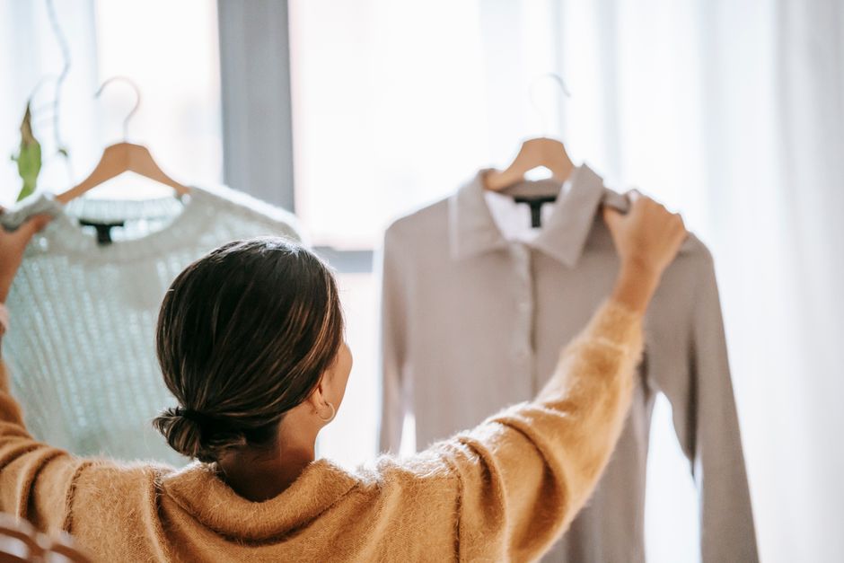 Back view of faceless female in casual clothes holding hangers with various garments while choosing outfit