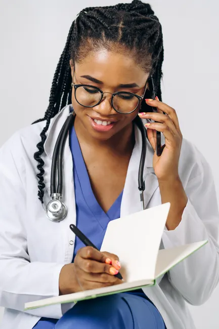 Black female doctor writing on a notebook while talking on a phone, wearing eyeglasses.