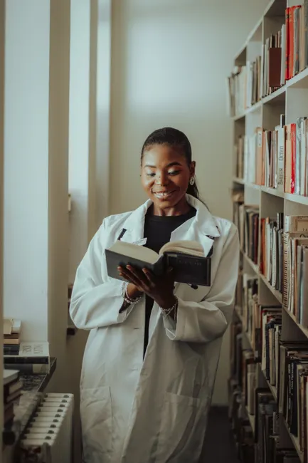 Black female scientist reading a book in a library, smiling, and wearing a white coat.