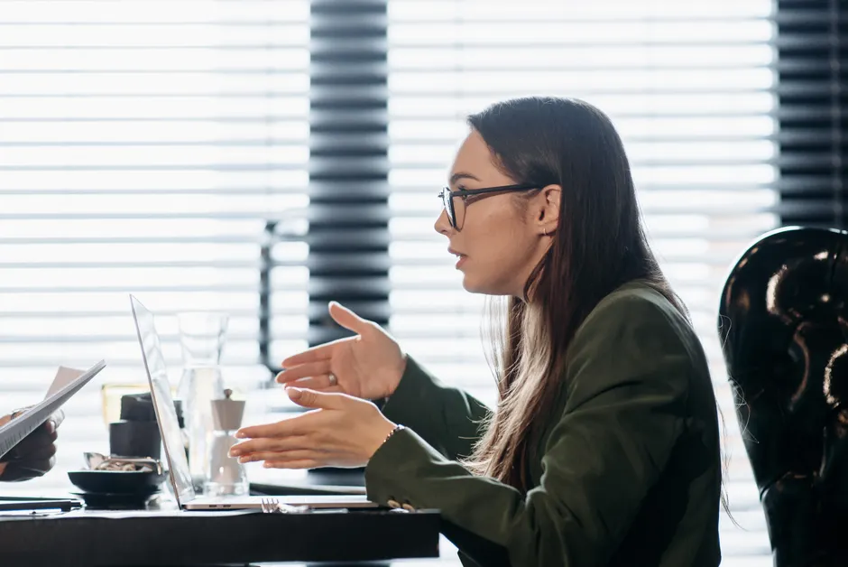 Caucasian woman with eyeglasses discussing and gesturing during a business meeting in a modern office.