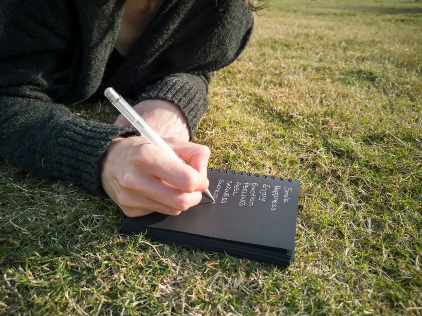 Close-up of a person writing emotions in a notebook while lying on grass outdoors.