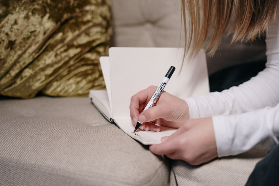 Close-up of a person writing in a notebook with a pen, sitting on a couch indoors.