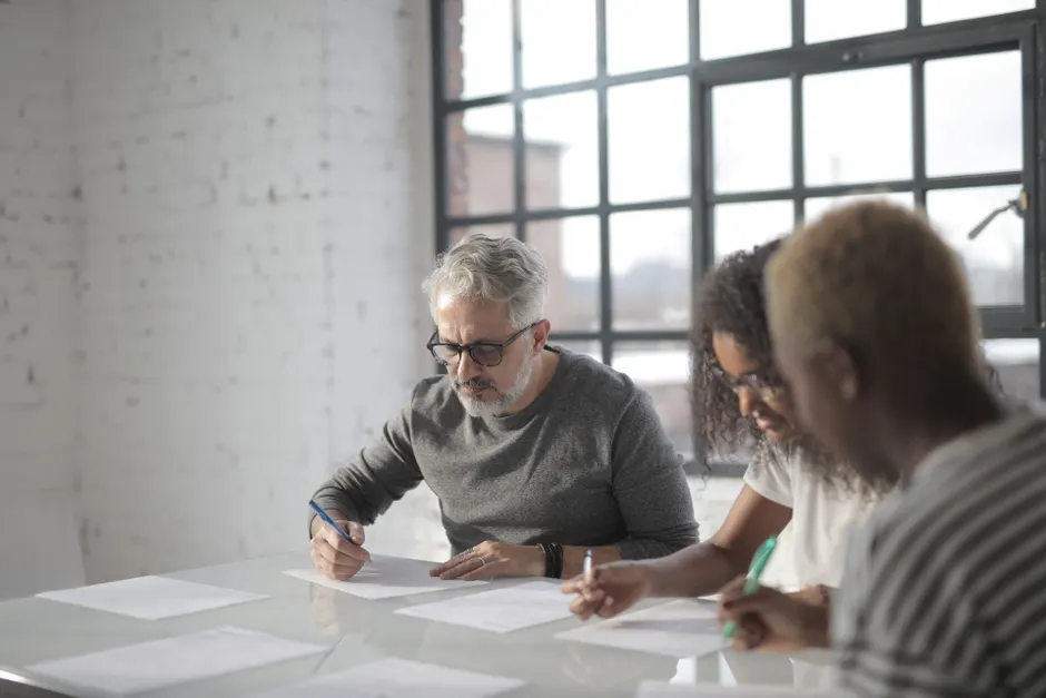 Concentrated multiracial people of different ages sitting at table near while writing on sheets of paper with pen in afternoon