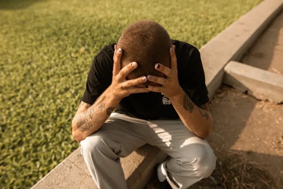 Distressed young man with tattoos sitting outdoors holding his head.