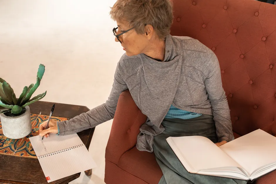 Elderly woman writing notes with a notebook while seated comfortably indoors.