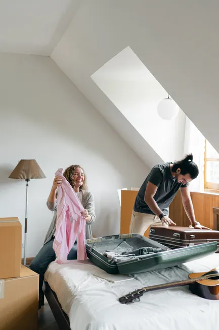 Full body happy young couple packing suitcases and carton boxes while preparing to move into new apartment in empty bedroom on sunny day